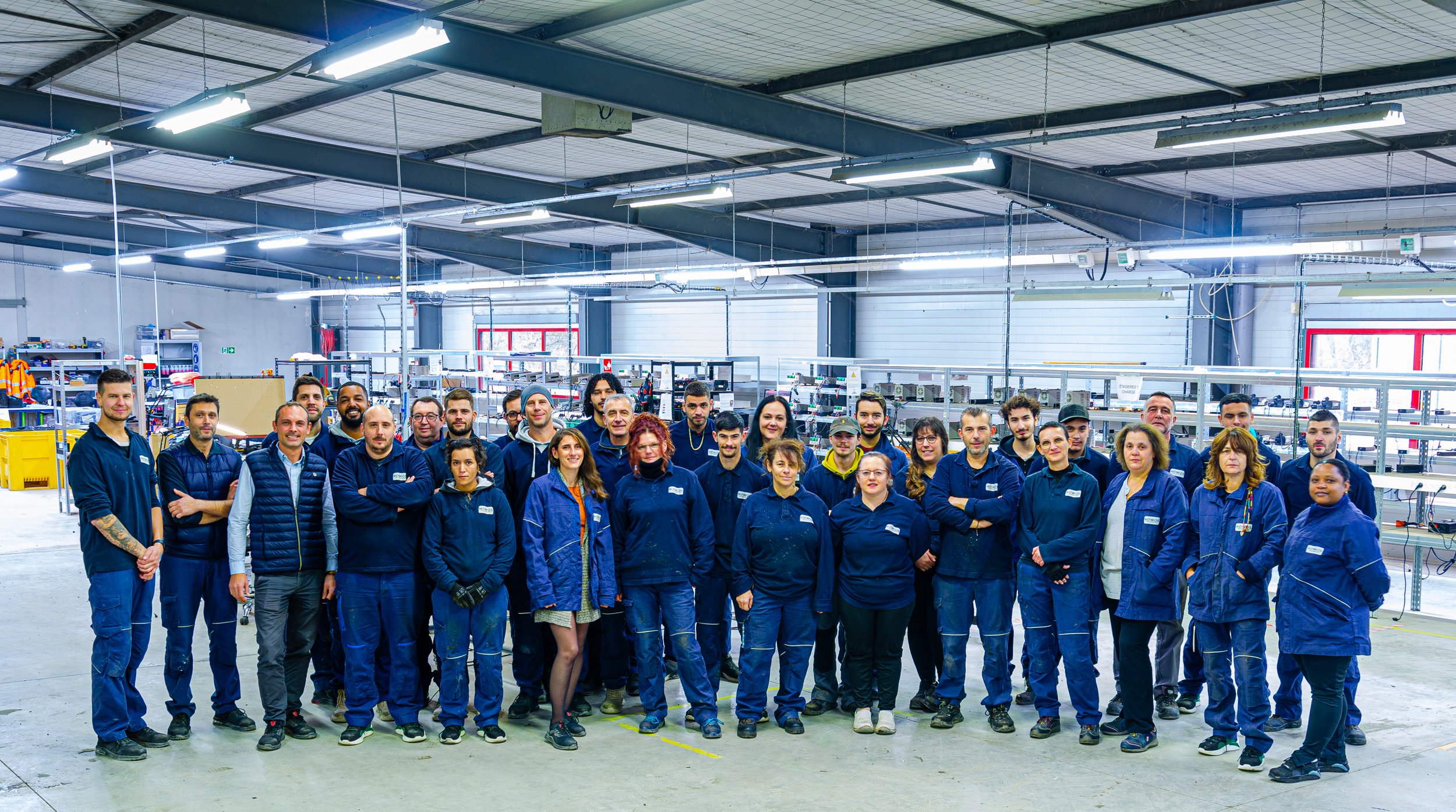 Group of factory workers standing inside a manufacturing plant with industrial equipment and shelving in the background.
