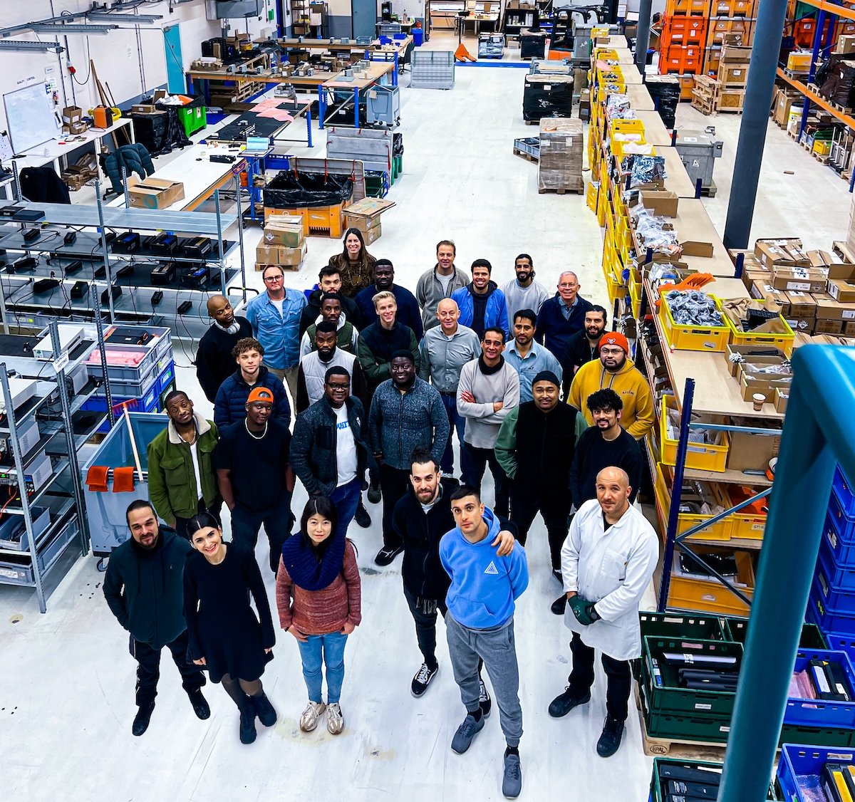 Group of diverse workers standing inside a warehouse or manufacturing facility surrounded by shelves filled with boxes and industrial equipment.